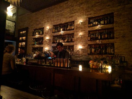A dimly lit bar with a bartender mixing drinks, shelves lined with bottles, and patrons seated at the counter.