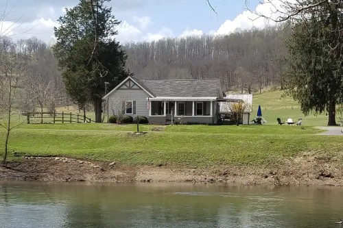 A cozy house by a calm river, surrounded by green grass and trees, with a scenic hillside in the background.
