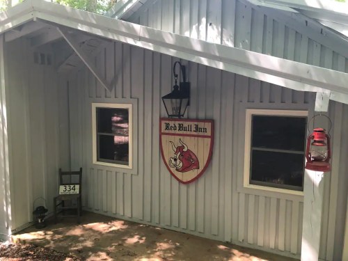 A rustic inn with a red bull sign, lantern, and a small chair outside, surrounded by trees.
