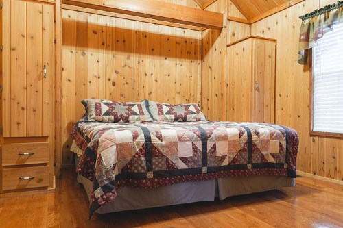 Cozy wooden bedroom featuring a quilted bedspread, wooden walls, and a window with curtains.