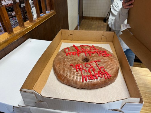 A large, round cake in a box with red icing writing on top, held by a person off to the side.