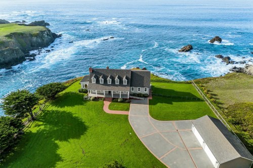 Aerial view of a coastal home with a large lawn, driveway, and ocean waves crashing against rocky cliffs nearby.