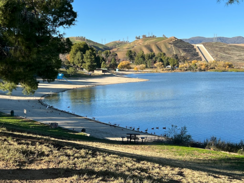 A serene lake surrounded by hills, with a sandy beach and trees, under a clear blue sky.