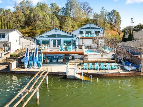 Aerial view of a lakeside property with two houses, a deck, kayaks, and seating by the water. Trees in the background.