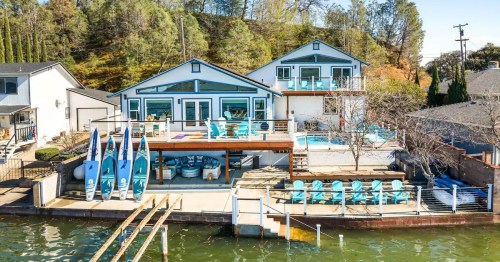 A lakeside property featuring two houses, a pool, and outdoor seating, surrounded by trees and water.