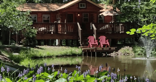 A cozy red cabin with a deck overlooking a pond, featuring two Adirondack chairs and surrounded by greenery and flowers.