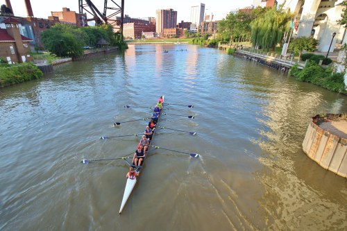 A rowing team in a boat glides through a calm river, surrounded by city buildings and greenery.