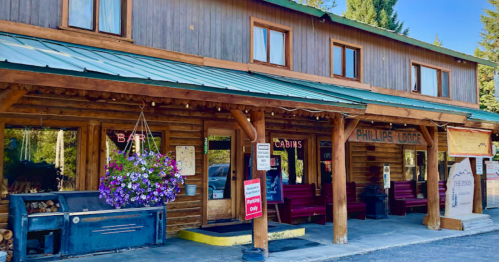 A rustic wooden lodge with a green roof, featuring a bar sign and flower baskets, surrounded by trees.