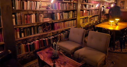 Cozy library setting with bookshelves, two armchairs, a wooden table, and warm lighting.