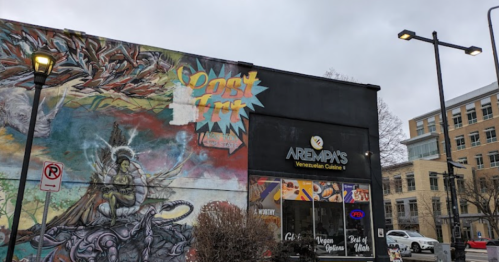 A colorful mural on a building next to a restaurant sign for Arepas Venezuelan Cuisine, with a cloudy sky above.