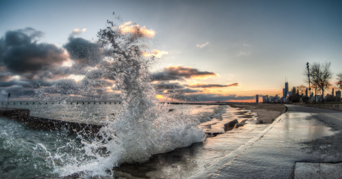 A wave crashes against a shoreline at sunset, with a city skyline in the background and dramatic clouds overhead.