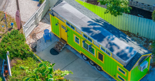 A colorful tiny house on wheels, featuring green and yellow siding, surrounded by greenery and a patio chair.