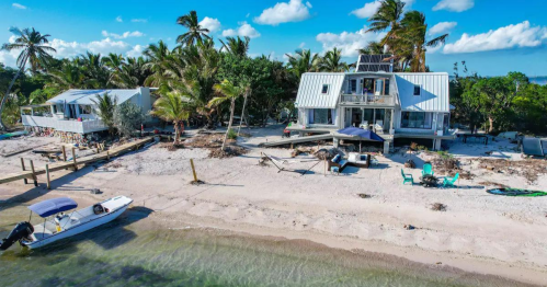 Aerial view of a beach house surrounded by palm trees, with a boat on the shore and clear blue skies.