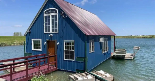 A blue houseboat with a red roof, situated on calm water, with a wooden dock and small boat nearby.
