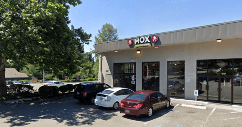 A storefront with a sign reading "MOX," featuring parked cars and trees in the background on a sunny day.