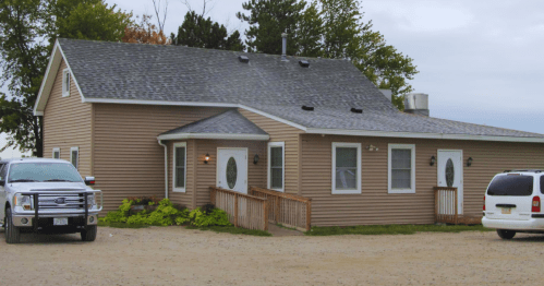 A beige house with a gray roof, surrounded by trees, and two parked vehicles in a gravel area.