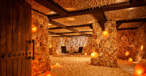 A tranquil salt room with stone walls, glowing salt lamps, and two chairs on sandy ground, creating a serene atmosphere.