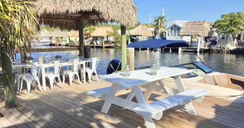 A waterfront deck with a picnic table, chairs, and thatched-roof huts overlooking a calm canal.