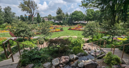 A lush garden with colorful flower beds, greenery, and a small stream, framed by trees under a partly cloudy sky.