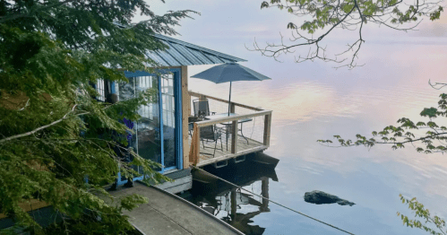A serene lakeside cabin with a deck, umbrella, and reflections on the water, surrounded by trees.