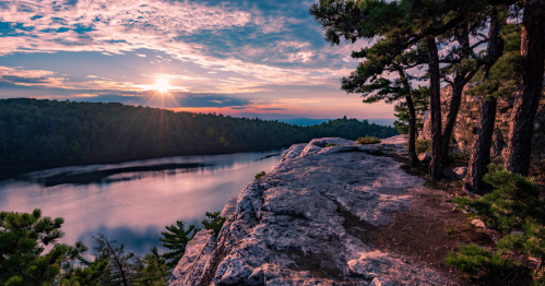 Sunset over a calm lake, viewed from a rocky cliff surrounded by trees, with colorful clouds in the sky.