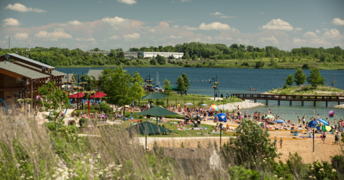 A sunny beach scene with people enjoying the water, green trees, and a distant building by the lake.