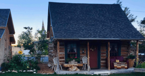 A cozy log cabin with a sloped roof, surrounded by greenery and featuring a small porch with chairs.