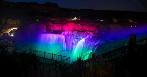 A vibrant waterfall illuminated in colorful lights at night, surrounded by rocky cliffs and a scenic landscape.