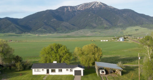 A white house with a black roof surrounded by green fields and trees, with mountains in the background.