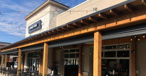 Exterior of a restaurant with wooden beams, large windows, and string lights, under a clear blue sky.