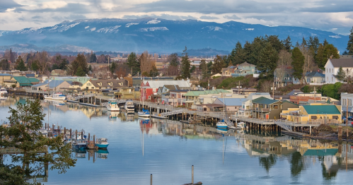 A scenic view of a waterfront town with colorful houses, boats, and mountains in the background under a cloudy sky.