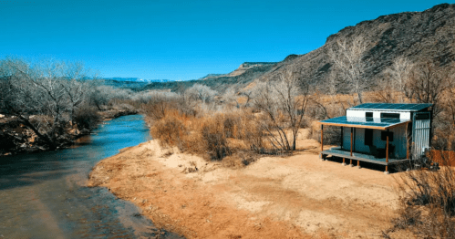 A small cabin by a river, surrounded by dry land and sparse trees, with mountains in the background under a clear blue sky.