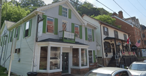 A charming two-story building with green shutters, next to another historic structure, set in a tree-lined street.