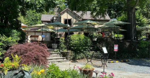 A charming restaurant with outdoor seating under green umbrellas, surrounded by lush greenery and colorful flowers.