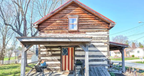 A rustic log cabin with a red roof and an American flag door, surrounded by trees and a small yard.