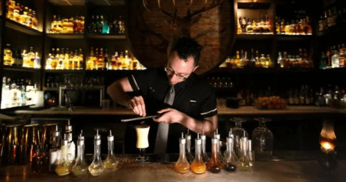 A bartender carefully prepares a cocktail at a well-stocked bar, surrounded by various bottles and glassware.