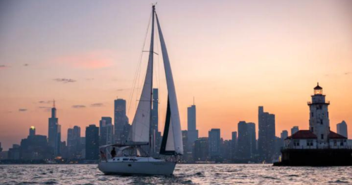 A sailboat glides on water at sunset, with a city skyline and lighthouse in the background.