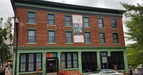 Historic brick building with green trim, featuring large windows and a banner, surrounded by trees and a street.