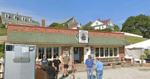 A brick pub with a sign, surrounded by people and houses on a sunny day. Outdoor seating is visible.