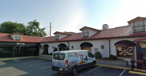 A restaurant with a red-tiled roof and a delivery van parked in front, surrounded by greenery and a clear sky.