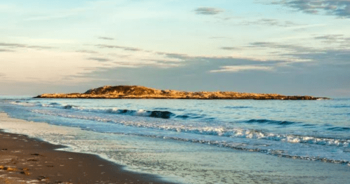 A serene beach scene with gentle waves lapping at the shore and a rocky island in the distance under a colorful sky.