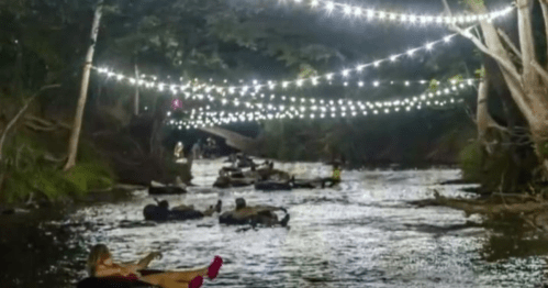 People floating on inner tubes in a river at night, illuminated by string lights above.