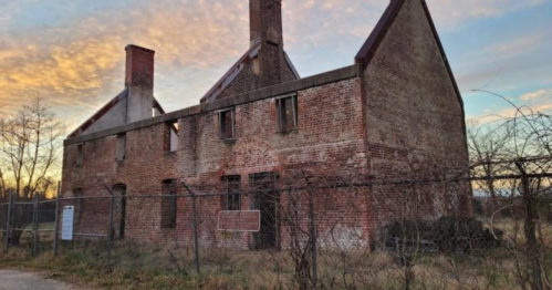 Abandoned brick building with a partially collapsed roof, surrounded by a fence and trees against a colorful sunset sky.