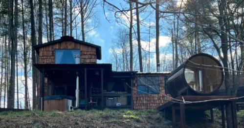 A rustic wooden cabin surrounded by trees, featuring a large round hot tub on a deck. Bright blue sky above.