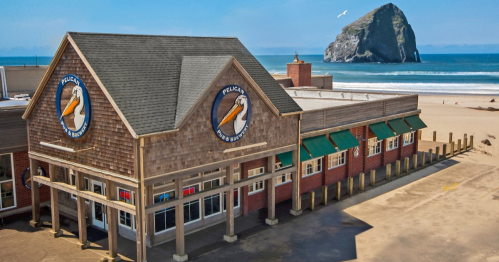 A beachside restaurant with a wooden exterior and large windows, overlooking the ocean and a rocky outcrop in the distance.