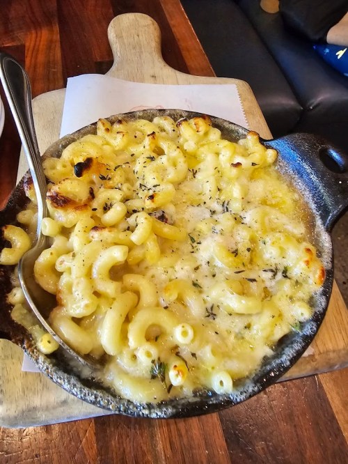 A baked macaroni and cheese dish in a cast iron skillet, topped with golden cheese and herbs, served on a wooden board.