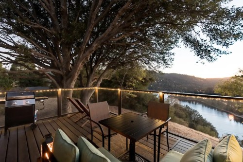 A cozy deck with a table and chairs overlooks a river and trees at sunset, featuring a grill and soft lighting.