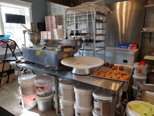 A bakery workspace with equipment, trays of donuts, and containers of ingredients on stainless steel tables.
