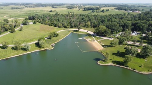 Aerial view of a serene lake surrounded by green fields and trees, with a small beach area and nearby camping spots.
