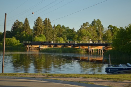 A wooden bridge over calm water, surrounded by trees and greenery, with a boat docked nearby under a clear sky.